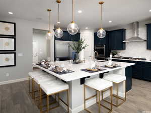 Kitchen featuring blue cabinets, a breakfast bar, a center island with sink, appliances with stainless steel finishes, and wall chimney range hood