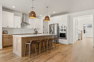 Kitchen featuring white cabinetry, appliances with stainless steel finishes, a kitchen island with sink, and wall chimney range hood