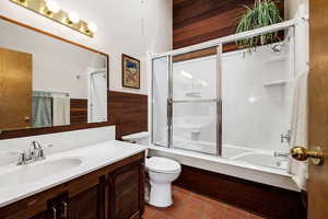 Full bathroom with wood walls, shower / bath combination with glass door, vanity, toilet, and tile patterned floors