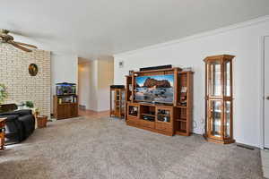 Carpeted living room featuring visible vents, ornamental molding, a ceiling fan, a textured ceiling, and brick wall