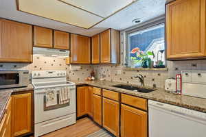 Kitchen with white appliances, light stone counters, brown cabinets, under cabinet range hood, and a sink