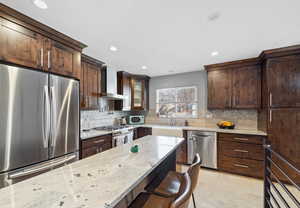 Kitchen with sink, backsplash, light stone counters, stainless steel appliances, and wall chimney range hood