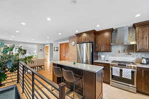 Kitchen featuring wall chimney range hood, a kitchen breakfast bar, stainless steel appliances, a center island, and light stone counters