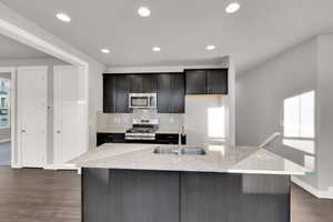 Kitchen featuring dark wood-type flooring, sink, appliances with stainless steel finishes, an island with sink, and light stone countertops