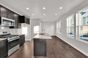 Kitchen with appliances with stainless steel finishes, dark hardwood / wood-style floors, light stone countertops, a kitchen island with sink, and decorative backsplash