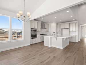 Kitchen featuring white cabinetry, hanging light fixtures, wall chimney exhaust hood, and a center island with sink