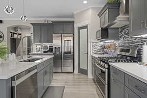 Kitchen with gray cabinetry, stainless steel appliances, hanging light fixtures, and wall chimney range hood