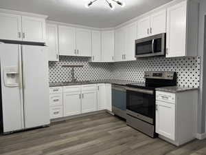 Kitchen with tasteful backsplash, appliances with stainless steel finishes, dark wood-type flooring, and white cabinets