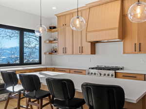 Kitchen with light brown cabinetry, hanging light fixtures, custom range hood, and stove