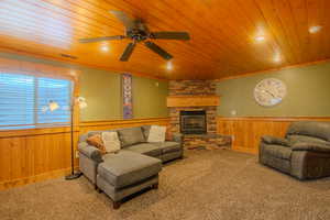 Carpeted living room featuring a stone fireplace, wooden ceiling, ceiling fan, and wood walls