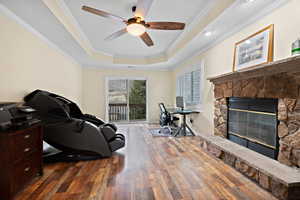 Office space featuring crown molding, dark wood-type flooring, ceiling fan, a tray ceiling, and a stone fireplace