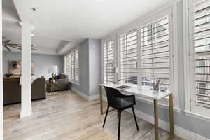 Home office featuring decorative columns, ceiling fan, a raised ceiling, a healthy amount of sunlight, and light hardwood / wood-style flooring