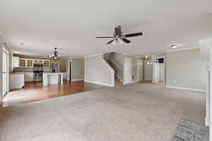 Unfurnished living room with light colored carpet, stairs, ornamental molding, ceiling fan with notable chandelier, and baseboards