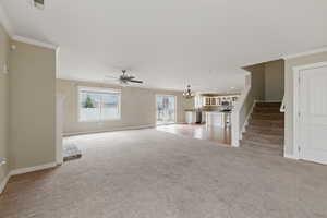 Unfurnished living room with stairs, crown molding, ceiling fan with notable chandelier, baseboards, and light colored carpet