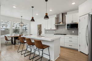 Kitchen with wall chimney range hood, sink, a kitchen island with sink, stainless steel appliances, and white cabinets