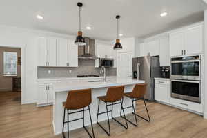 Kitchen with wall chimney exhaust hood, sink, white cabinetry, hanging light fixtures, and stainless steel appliances