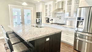 Kitchen featuring a center island with sink, appliances with stainless steel finishes, wall chimney range hood, and white cabinets