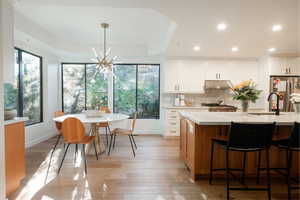 Kitchen featuring stainless steel fridge, an island with sink, white cabinets, decorative light fixtures, and light wood-type flooring