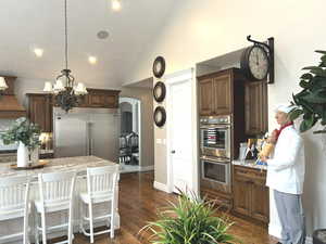 Kitchen with premium range hood, dark brown cabinetry, hanging light fixtures, appliances with stainless steel finishes, and dark hardwood / wood-style flooring