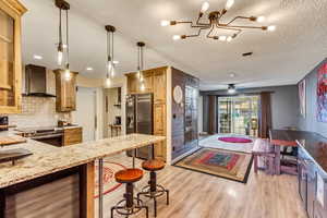 Kitchen featuring decorative light fixtures, tasteful backsplash, stainless steel appliances, wall chimney range hood, and light hardwood / wood-style flooring