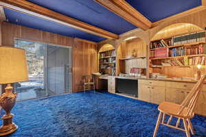 Carpeted home office featuring built in shelves, wood walls, built in desk, ornamental molding, and beamed ceiling