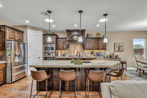 Kitchen featuring pendant lighting, a kitchen island with sink, stainless steel appliances, dark brown cabinetry, and custom range hood