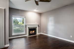 Unfurnished living room with a stone fireplace, dark wood-type flooring, and ceiling fan