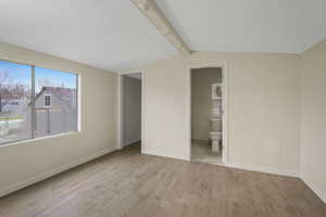 Unfurnished bedroom with vaulted ceiling with beams, ensuite bathroom, and light wood-type flooring