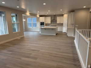 Kitchen with wall chimney range hood, a kitchen island with sink, white cabinetry, decorative backsplash, and stainless steel oven