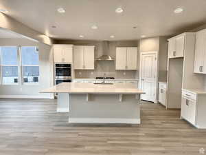 Kitchen featuring white cabinetry, stainless steel double oven, light stone countertops, a center island with sink, and wall chimney exhaust hood