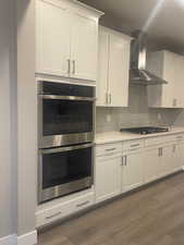 Kitchen featuring wall chimney range hood, dark hardwood / wood-style floors, white cabinets, decorative backsplash, and stainless steel double oven