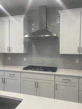 Kitchen featuring stainless steel gas stovetop, white cabinetry, decorative backsplash, light stone counters, and wall chimney range hood