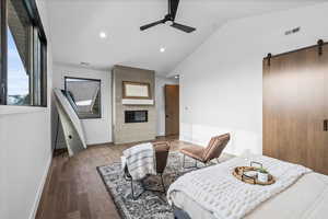 1st primary bedroom with lofted ceiling, a large fireplace, hardwood flooring, ceiling fan, and a barn door to bathroom.