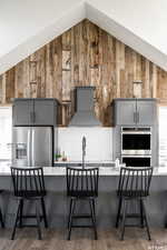 Kitchen featuring vaulted ceiling, appliances with stainless steel finishes, a kitchen breakfast bar, gray cabinets, and custom range hood