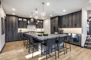 Kitchen featuring decorative light fixtures, dark brown cabinets, an island with sink, beverage cooler, and light hardwood / wood-style floors