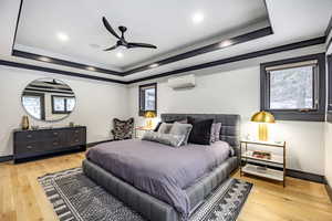 Bedroom with a tray ceiling, a wall unit AC, ceiling fan, and light wood-type flooring