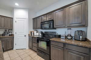 Kitchen featuring dark brown cabinetry, light tile patterned floors, black appliances, and dark stone countertops