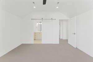 Unfurnished bedroom featuring lofted ceiling, ceiling fan, ensuite bathroom, light carpet, and a barn door