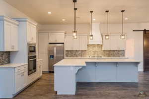 Kitchen with decorative light fixtures, appliances with stainless steel finishes, custom range hood, a kitchen island with sink, and white cabinets