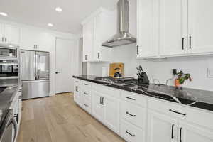 Kitchen featuring wall chimney exhaust hood, light hardwood / wood-style flooring, appliances with stainless steel finishes, dark stone counters, and white cabinets