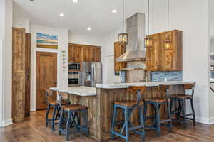 Kitchen with appliances with stainless steel finishes, decorative light fixtures, tasteful backsplash, range hood, and dark wood-type flooring