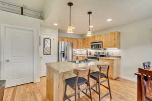 Kitchen with stainless steel appliances, light hardwood / wood-style floors, a center island with sink, decorative light fixtures, and light brown cabinets