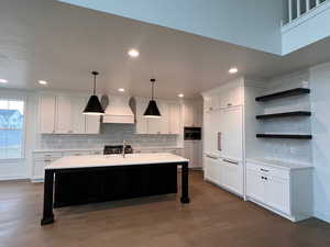 Kitchen with hanging light fixtures, custom range hood, white cabinets, a center island with sink, and stainless steel oven