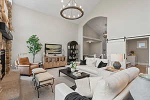 Living room featuring an inviting chandelier, high vaulted ceiling, carpet floors, a stone fireplace, and a barn door