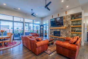 Living room with a stone fireplace, dark wood-type flooring, ceiling fan, and plenty of natural light