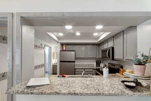 Kitchen featuring gray cabinetry, appliances with stainless steel finishes, kitchen peninsula, a raised ceiling, and light stone countertops