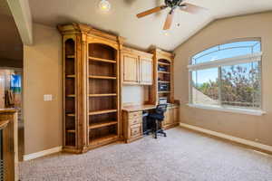 Unfurnished office featuring lofted ceiling, built in desk, light colored carpet, and ceiling fan