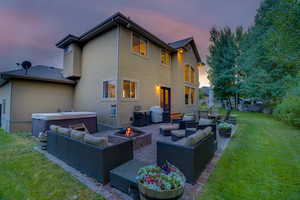 Back house at dusk featuring an outdoor living space with a fire pit, a lawn, a hot tub, and a patio