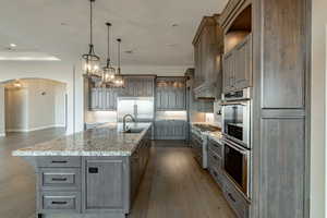 Kitchen with sink, a large island with sink, hanging light fixtures, light stone counters, and stainless steel appliances