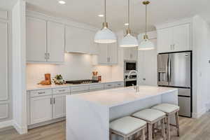 Kitchen featuring white cabinetry, hanging light fixtures, stainless steel appliances, a center island with sink, and light wood-type flooring